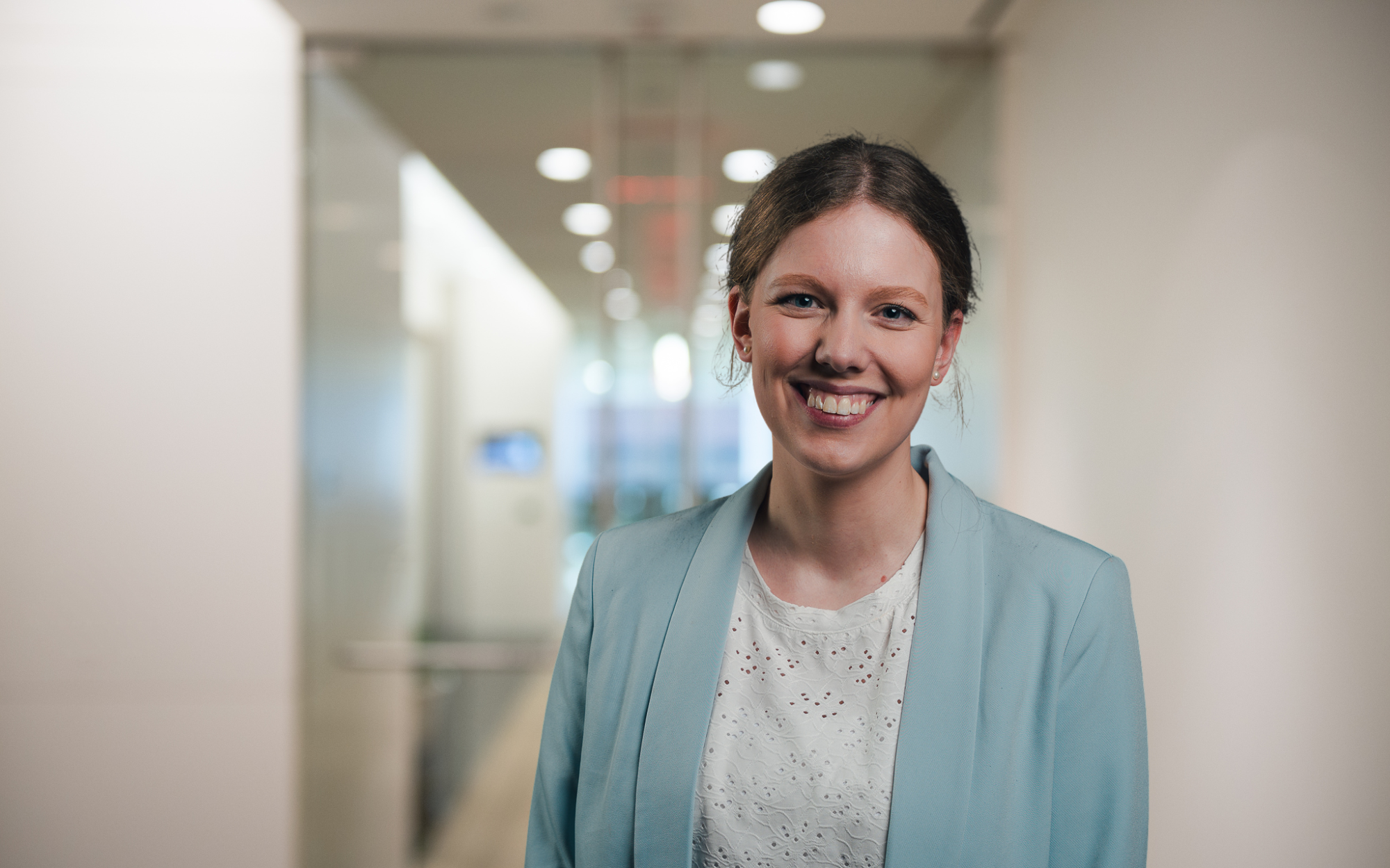 Porträtfoto von Lena Leitow, Mitarbeiterin im Bereich Marketing bei AUDIOSERVICE plus. Sie steht lächelnd in einem hellen Büroflur, trägt eine weiße Bluse und einen hellblauen Blazer.