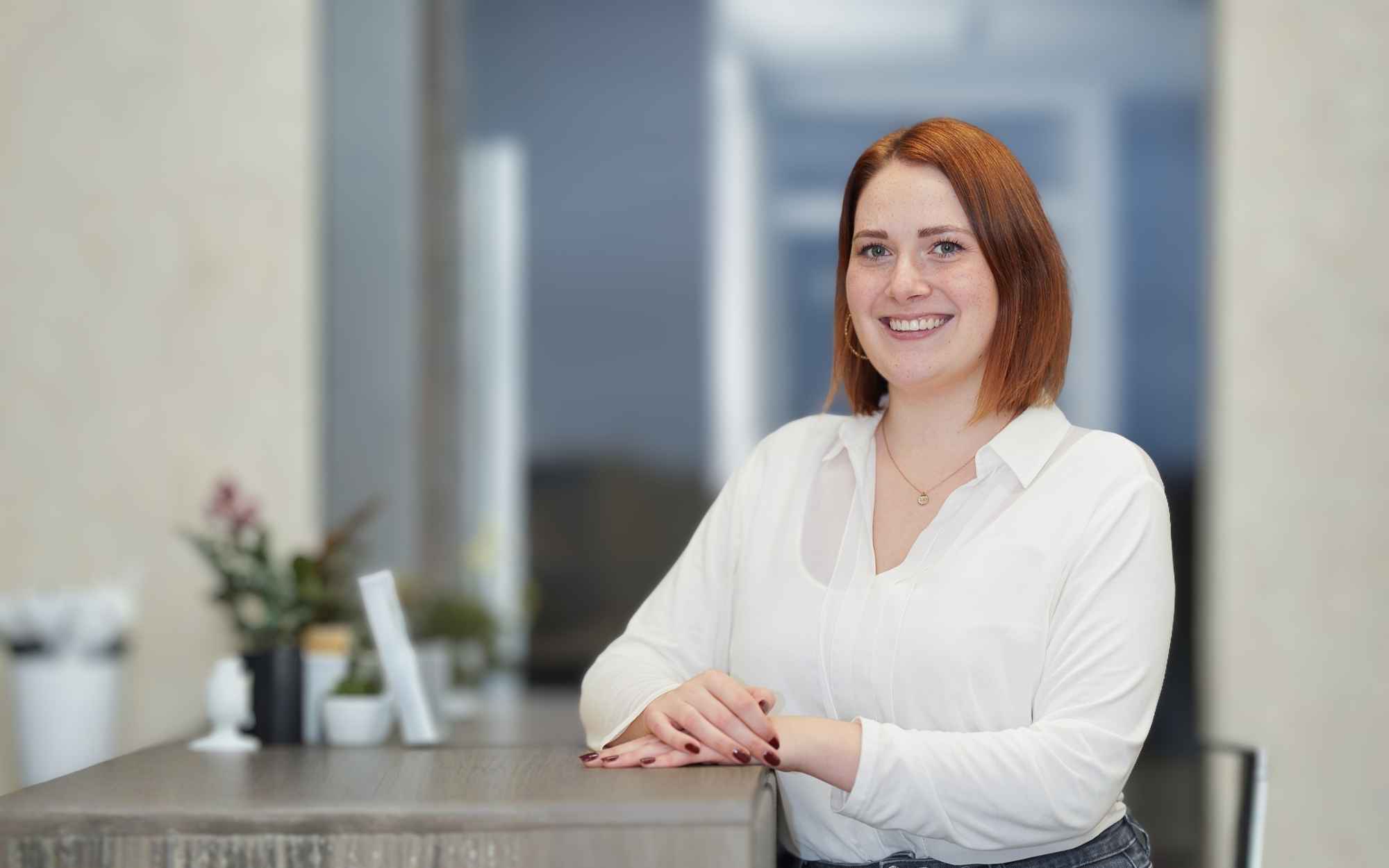 Porträt von Svenja Jesse: Die Online-Managerin von AUDIOSERVICE plus lehnt lächelnd an einem Tresen in einer hellen Büroumgebung.