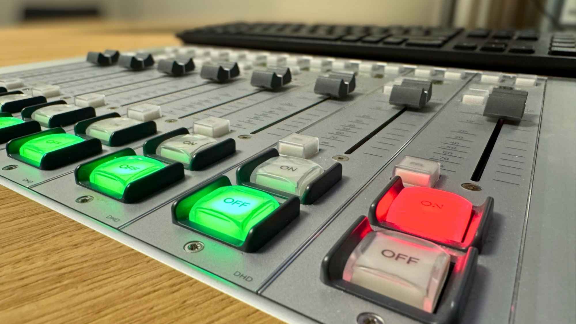 Nahaufnahme eines Radiostudio-Mischpults mit leuchtenden grünen und roten On-Off-Tasten sowie Reglern für Audiotechnik und Radio-Produktion.